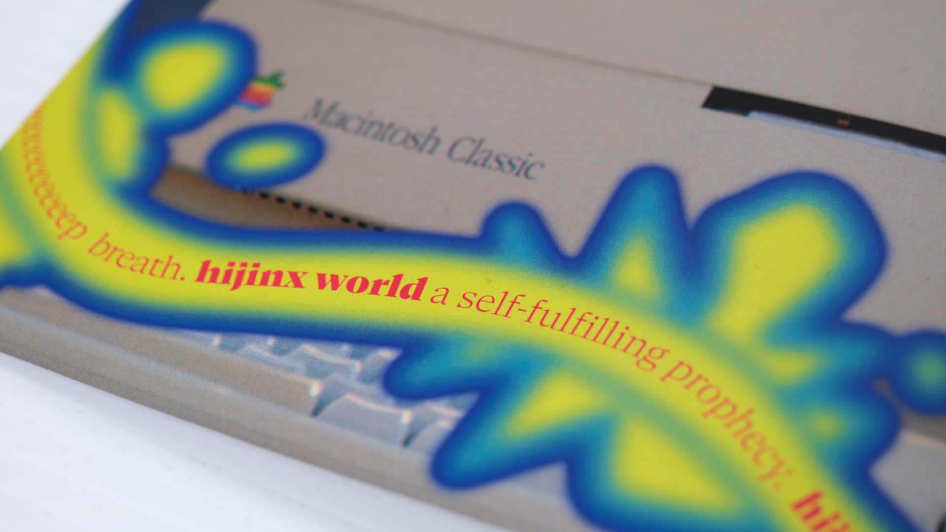 Close up of a magazine page with wavy text on a splotchy background overtop the corner of a Macintosh Classic that reads "deeeeeeep breath, hijinx world is a self-fulfilling prophecy"