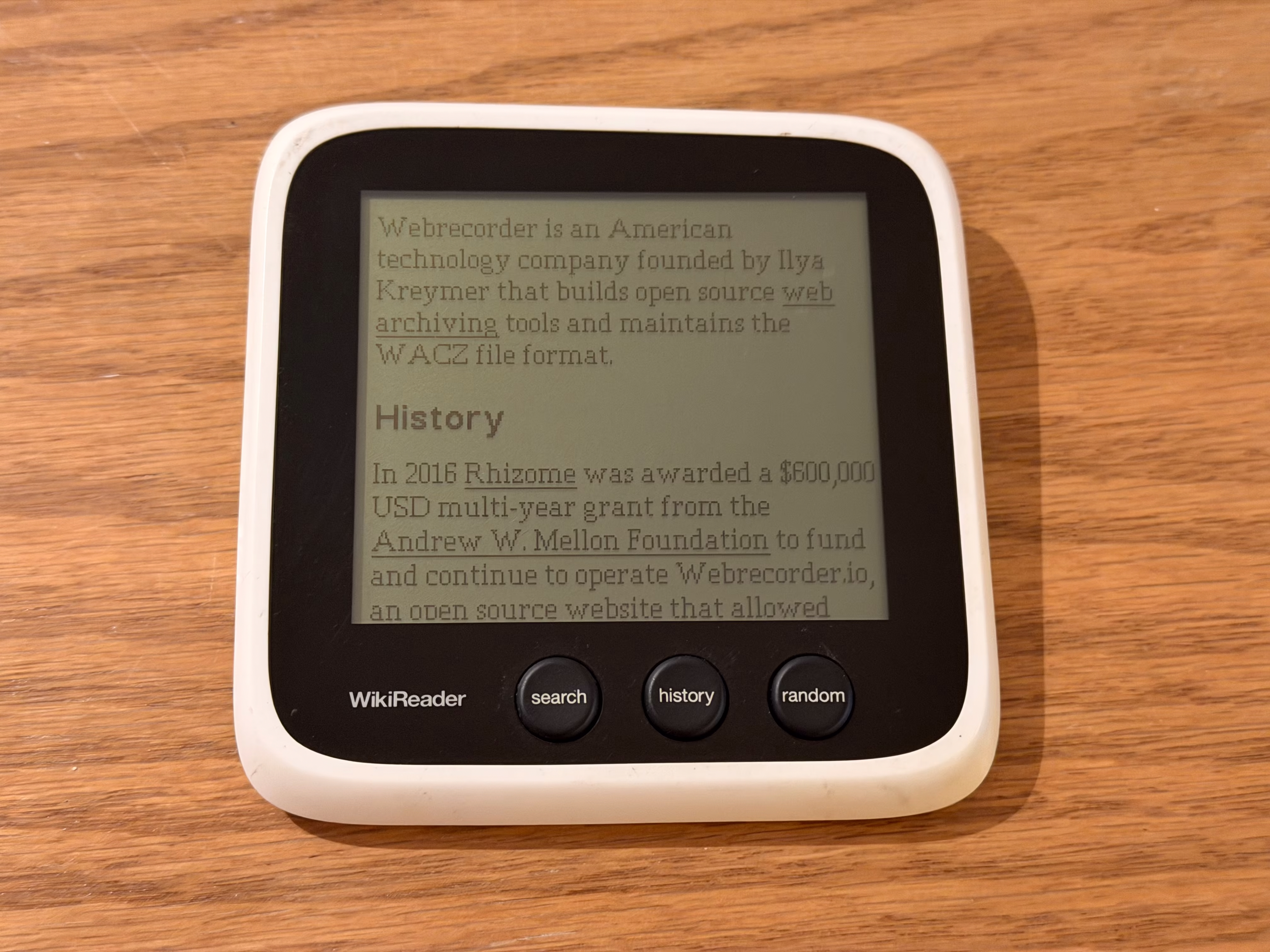 A square-ish device with comfortably rounded corners, large black bezels, and a monochromatic screen displaying a Wikipedia article. Three buttons are on the front labeled search, history, and random.