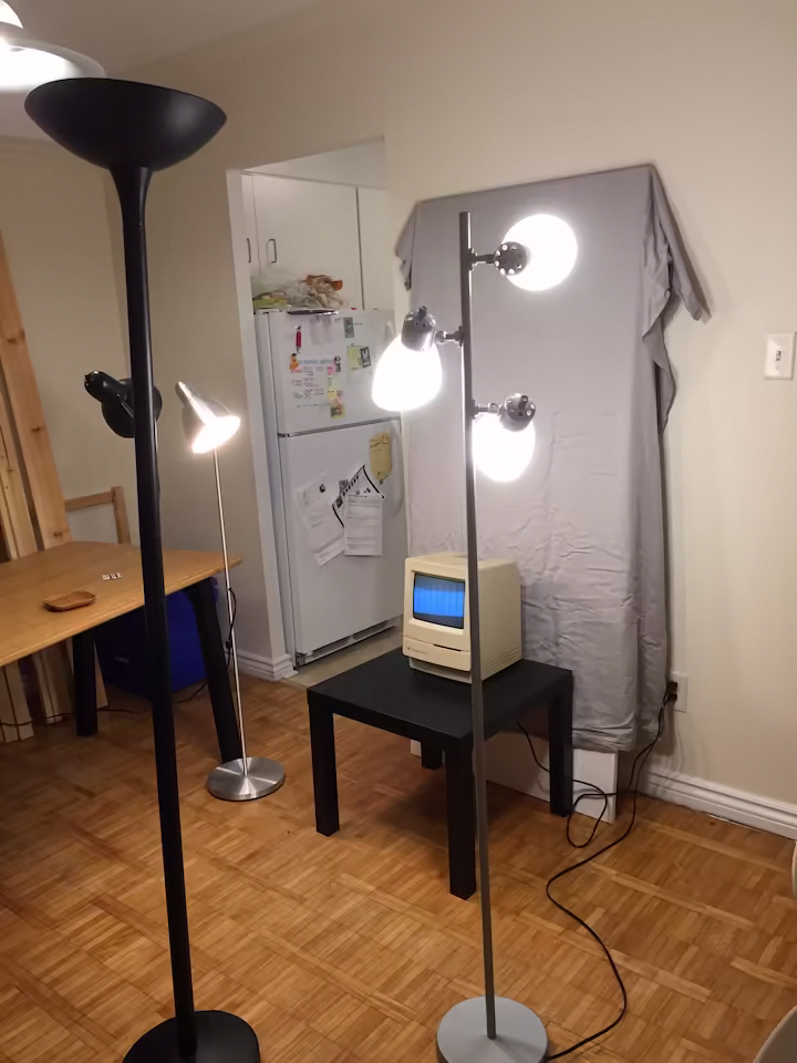 A Macintosh Classic set up in a living room with lights around it and a bedsheet as a backdrop behind it